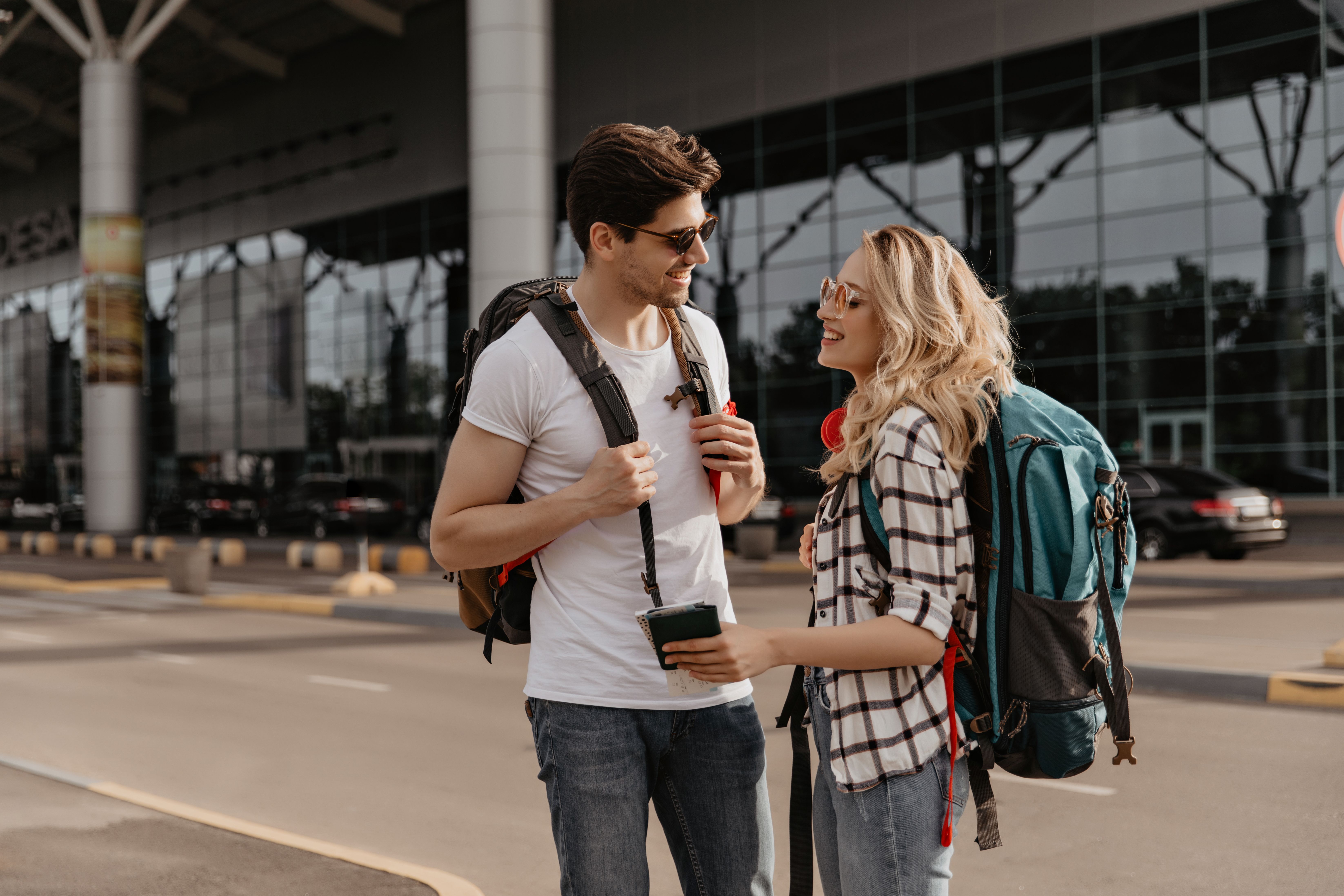 woman-plaid-shirt-man-white-tee-poses-with-backpacks-near-airport-travelers-sunglasses-talking-holding-passports.jpg